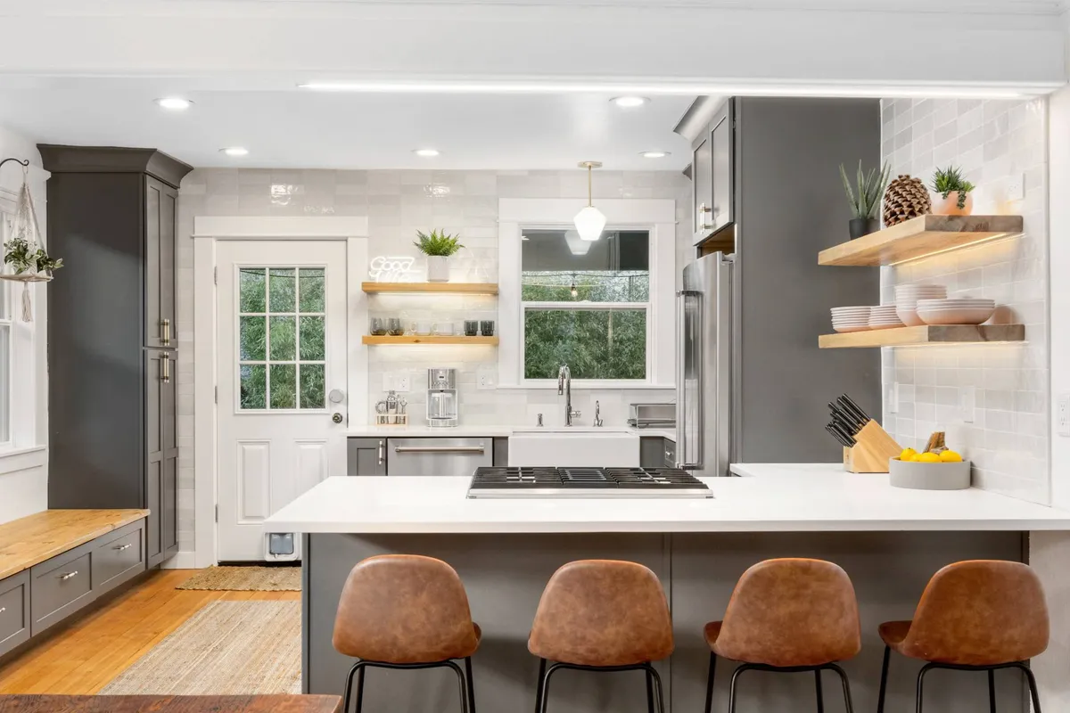 A bright, modern kitchen with a central island and bar stools, open shelving adorned with plants, and a welcoming layout for cooking and entertaining. The atmosphere is both inviting and contemporary, encouraging shared meals and social connection.