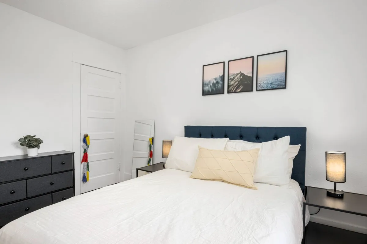 A serene bedroom featuring a blue headboard, three framed landscape pictures above the bed, nightstands with lamps, and a dresser with greenery. The room is bathed in natural light, creating a calm and inviting atmosphere.