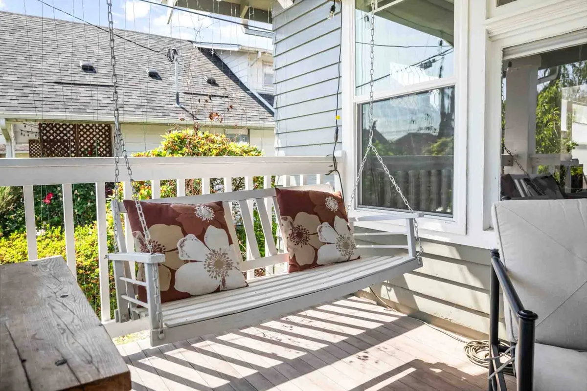 This image features a cozy swing with decorative pillows set near a large window, with a wooden table nearby. The view through the window hints at the outdoors, suggesting a spot perfect for enjoying a cup of coffee, relaxing during pleasant weather, or simply soaking in seasonal sunlight.