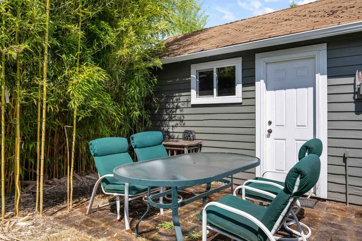 A tranquil outdoor patio framed by bamboo plants, with a table and chairs near the entrance door. The scene evokes a sense of relaxed socializing and the possibility of enjoying meals or coffee outdoors, connecting the home base to its lively urban environment.
