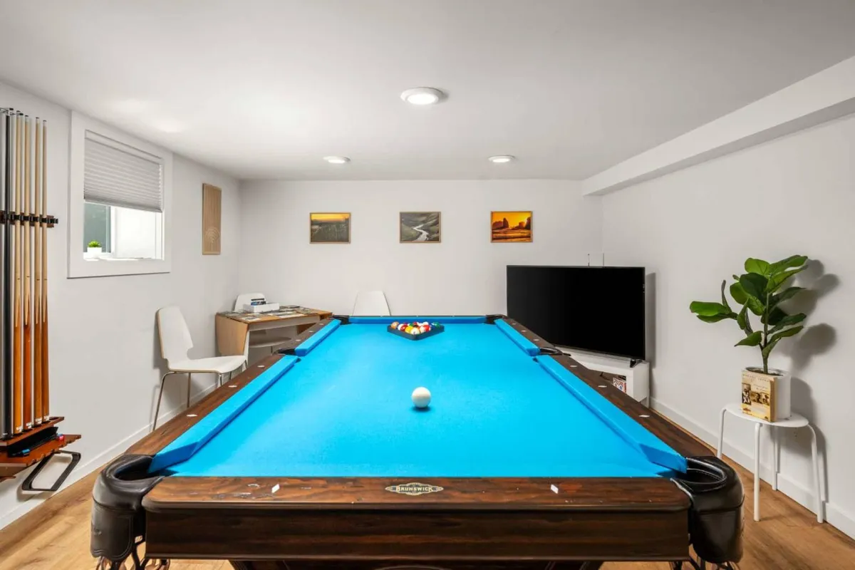 A stylish, leisurely interior space centered around a modern pool table, with a large TV, potted plant, and artwork on the wall. The mood is fun and social, ideal for game nights or gatherings.