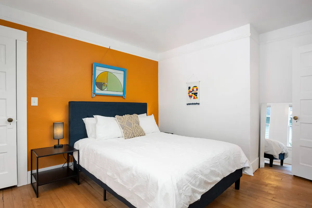 A comfortable bedroom with a bed against an orange accent wall, decorative artwork, a nightstand, and a mirror. The room is softly lit and appears welcoming and serene, ideal for restful nights and private moments.