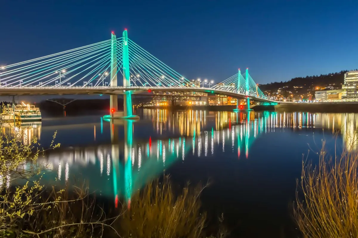 An illuminated bridge over the water at night, city lights reflecting on the river. The scene suggests nighttime activity and the liveliness of the local nightlife, as well as opportunities for riverside strolls and photography.