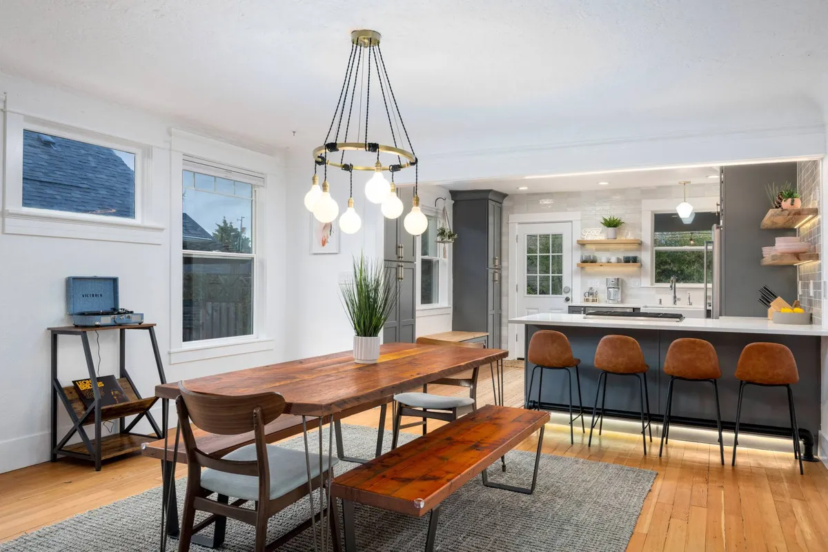 This image features a warm and inviting dining area with a prominent dining table, a kitchen bar adorned with stools, an elegant chandelier overhead, and large windows that fill the space with natural light. The open setup allows for a seamless flow between social dining and culinary preparation.