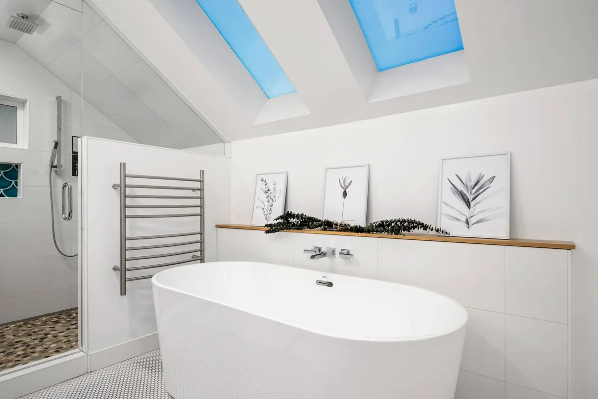 A spa-like primary bathroom filled with natural light from skylights, featuring a freestanding soaking tub, a heated towel rack, and tranquil framed botanical prints. The soothing atmosphere is accentuated by the warm glow of sunset visible through the skylights, evoking a relaxing, resort-style retreat.